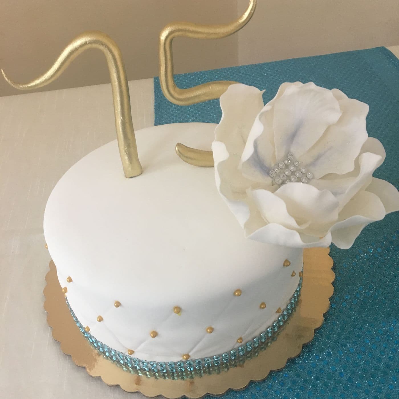 75th birthday cake in white fondant with a gold hand-made number topper, a large white sugar peony, teal crystal banding and gold dragée dots