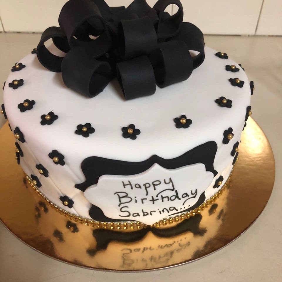Birthday cake in white fondant with a black fondant loop bow, small black flowers with gold centres and a hand-piped name plaque