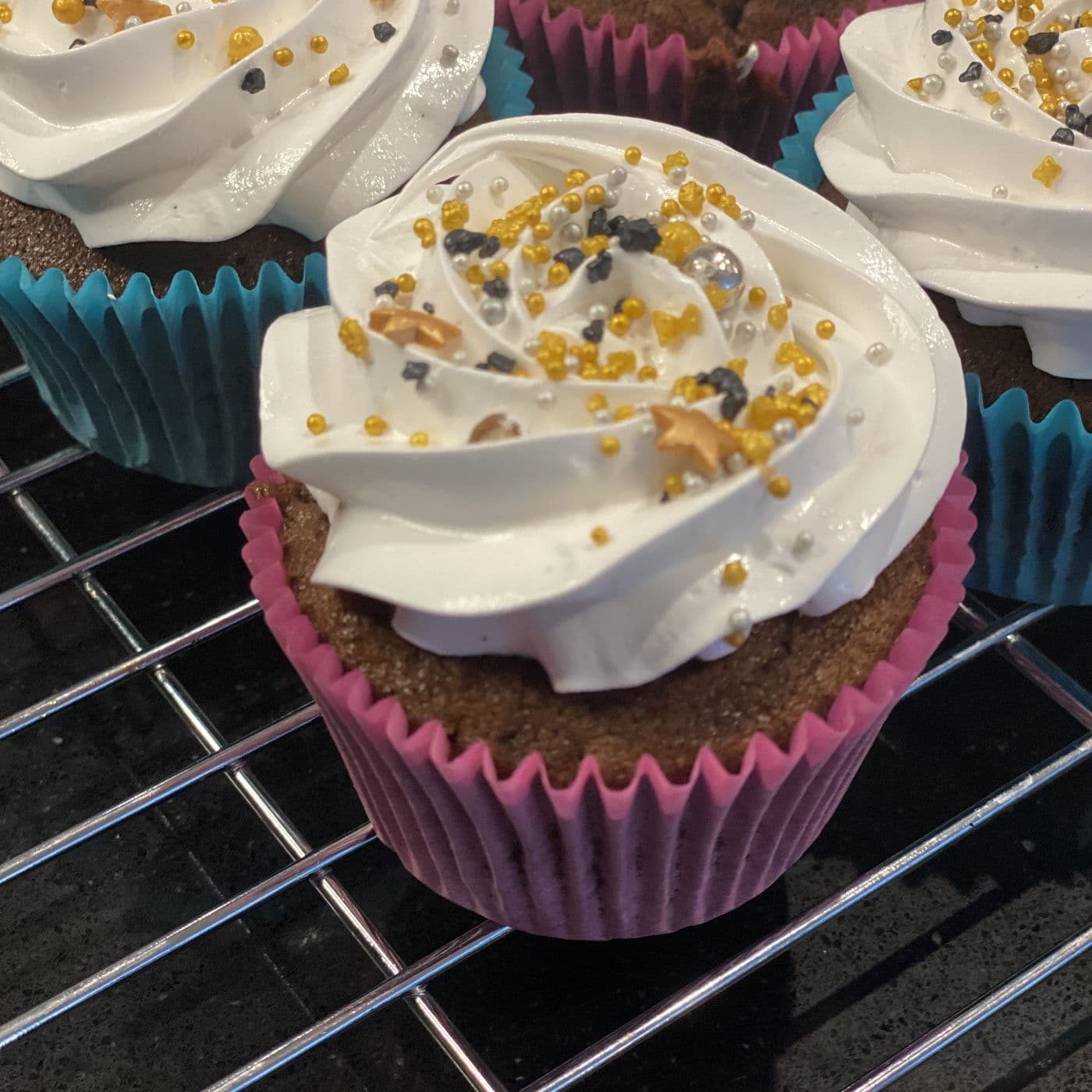 Close view of a chocolate cupcake with a tall piped buttercream swirl, gold and silver sprinkles, in a pink case