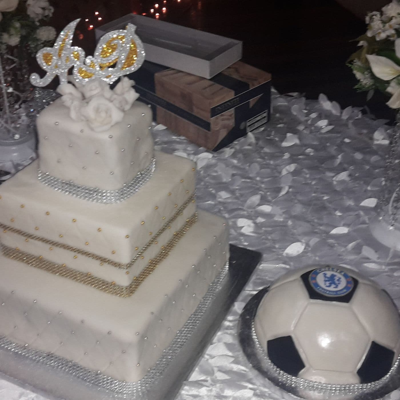 Three-tier square wedding cake in white fondant with silver and gold crystal banding, a monogram topper and hand-made sugar roses, set up at the venue beside a football-shaped groom's cake