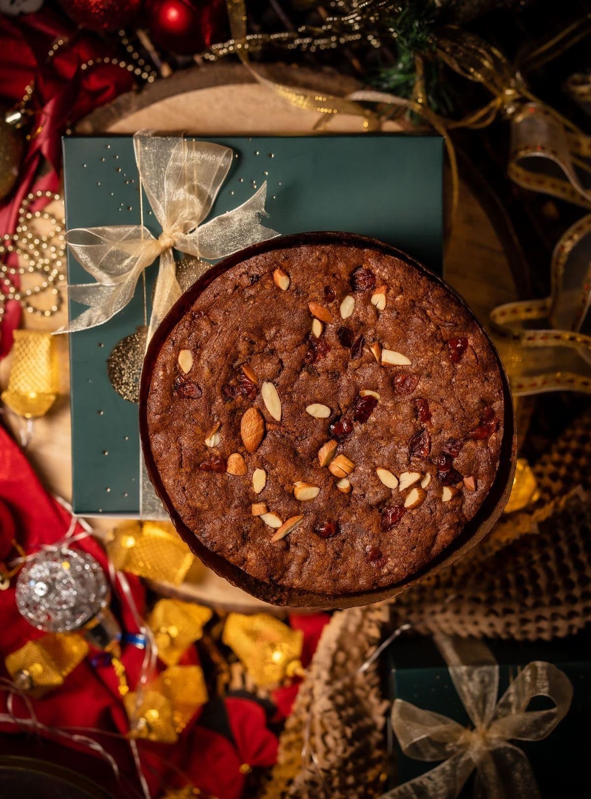 A dark, un-iced Christmas fruit cake beside a green gift box tied with gold ribbon