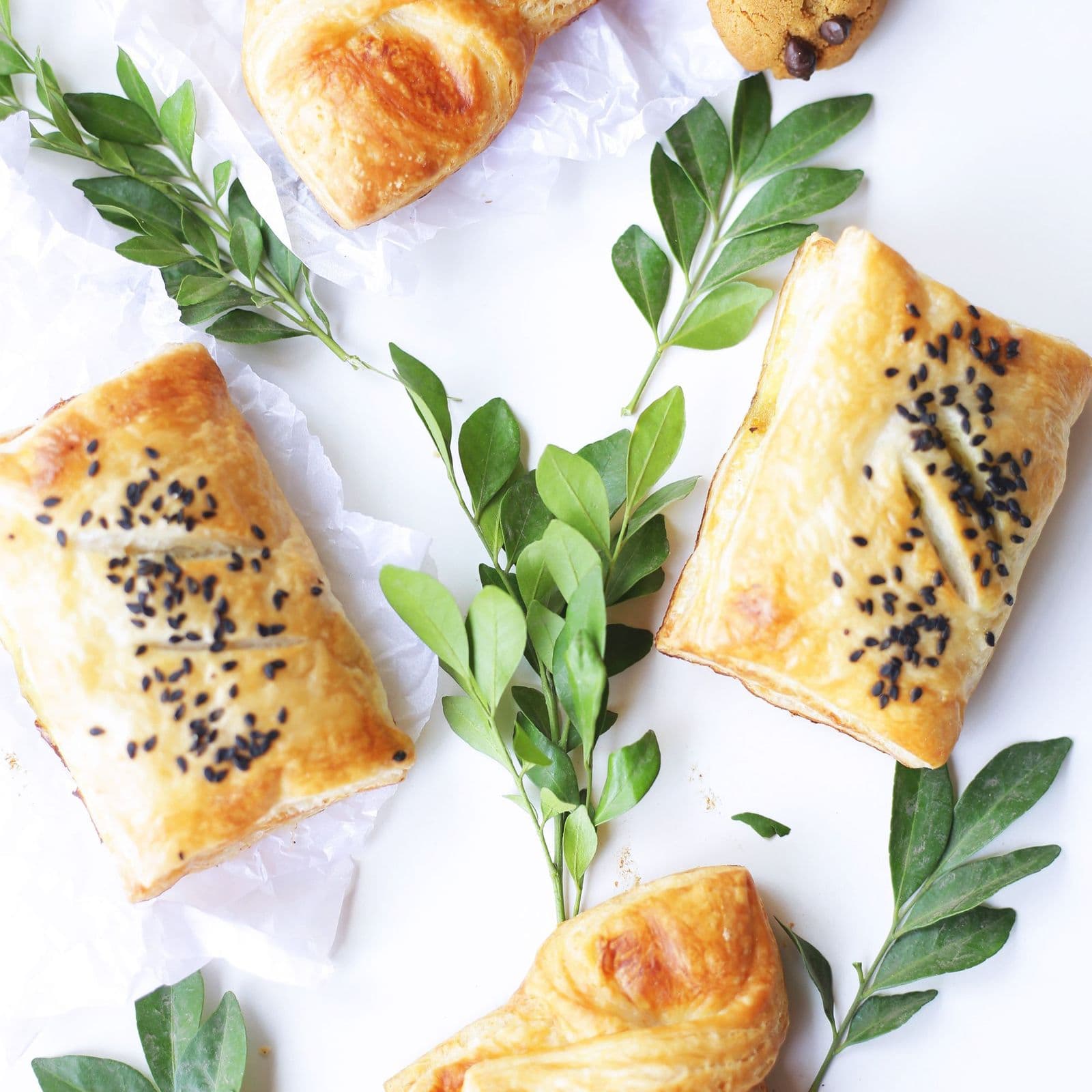 Golden puff pastry rolls scattered with black sesame, on parchment with fresh herbs