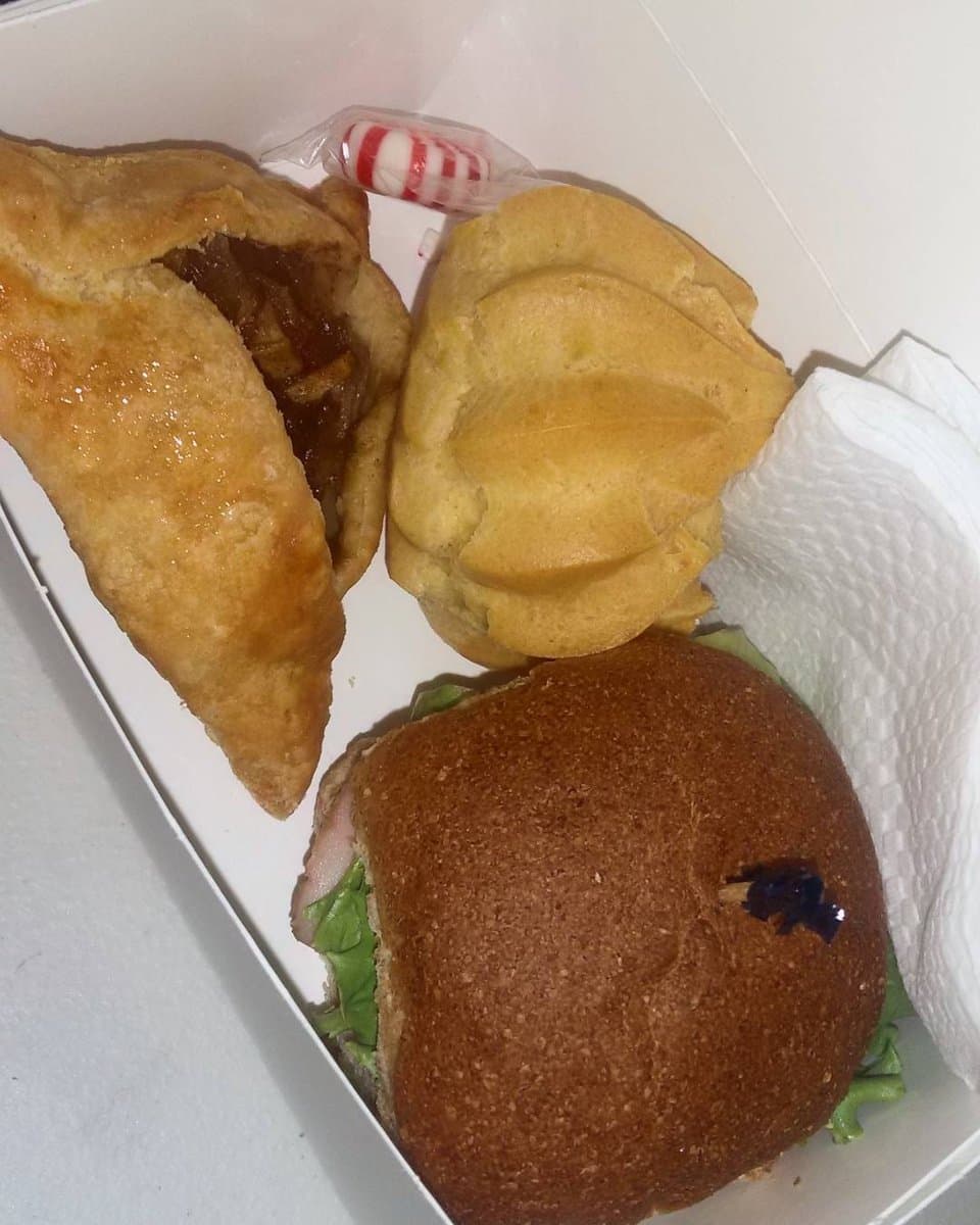 A white lunch box with a meat-filled pastry turnover, a choux puff, a wholemeal roll with lettuce and a striped sweet