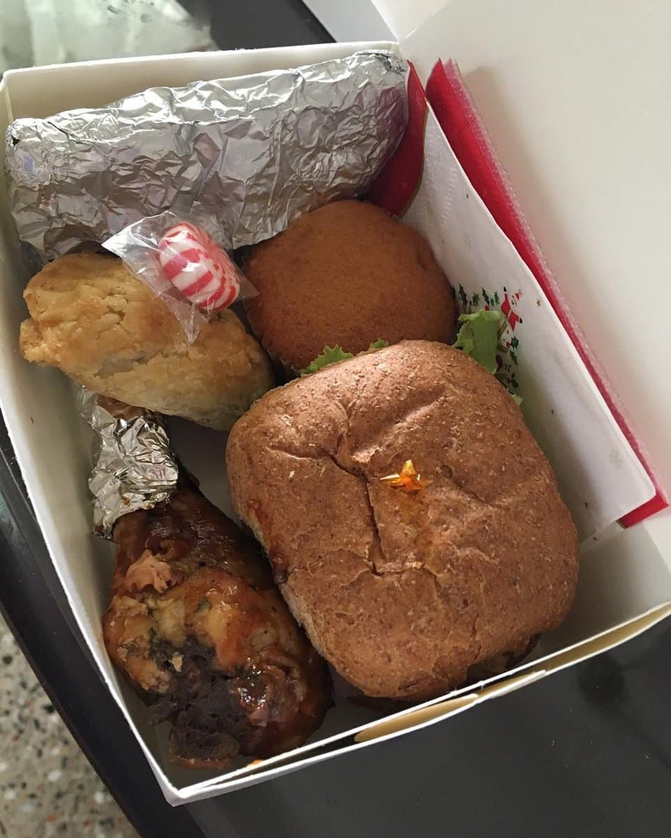 A lunch box with a wholemeal roll, a glazed chicken drumstick, a golden pastry, a plain cupcake and a striped sweet