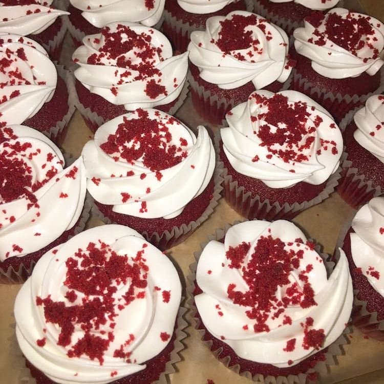 A box of red velvet cupcakes with white frosting rose swirls topped with red velvet crumbs