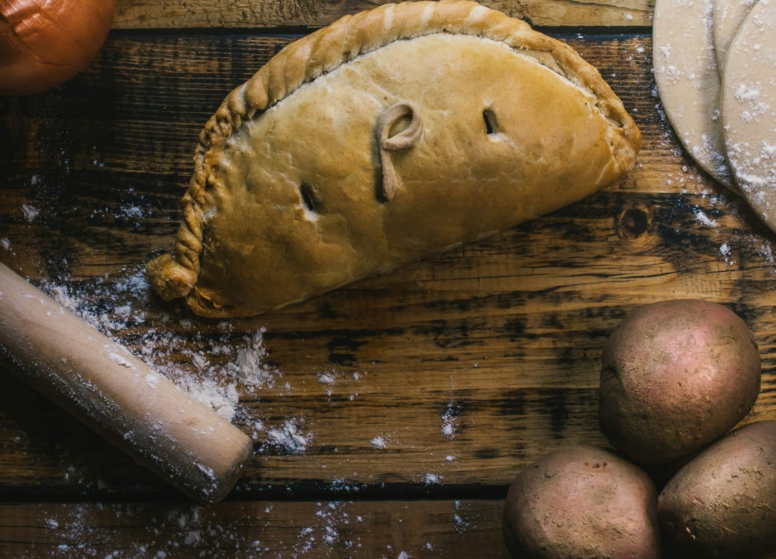 A golden, crimped half-moon pie on a floured wooden table beside potatoes and a rolling pin
